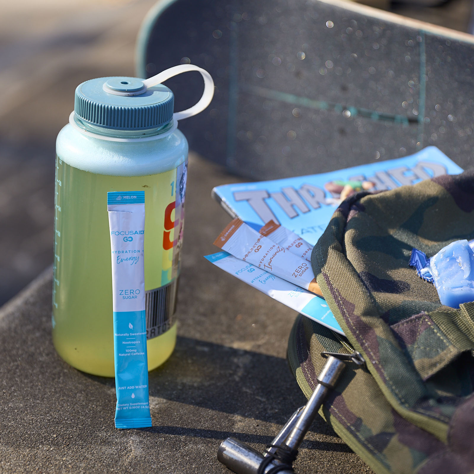 A water bottle with Focusaid GO beside it next to a skateboard, a magazine, and a backpack.