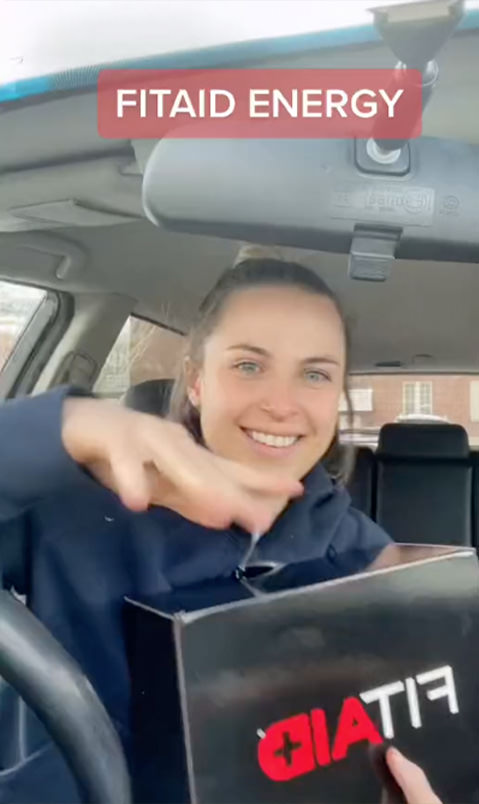 Brand ambassador in blue sweatshirt, sitting in car, holding a black FITAID box, read text across the top of the image reading "FITAID ENERGY"